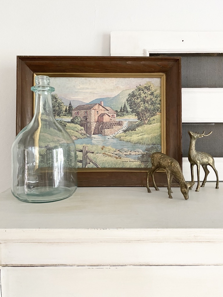 A framed vintage painting of an old mill with a vintage blue glass bottle and a pair of brass deer