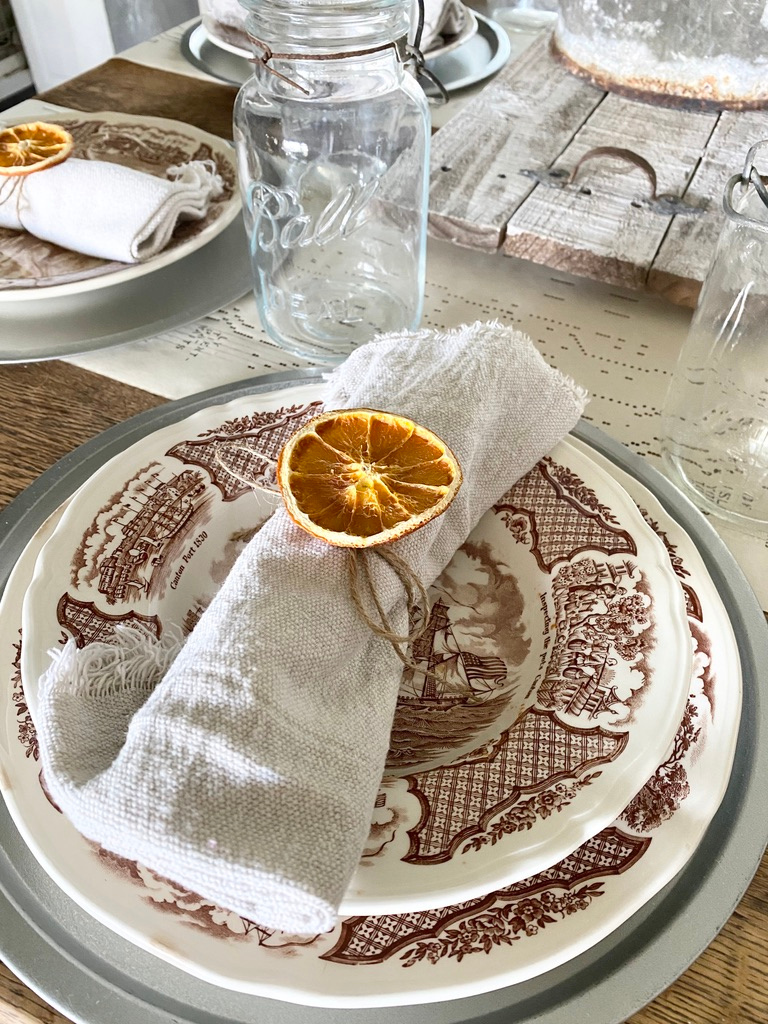 A brown ironstone transferware place setting with a dropcloth napkin and dried orange napkin ring